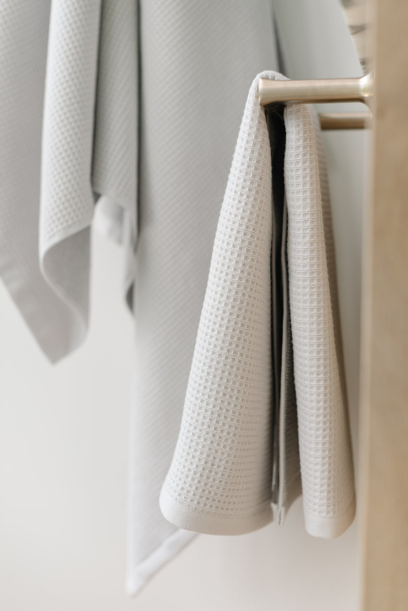 Light Grey Waffle Hang Towel handing from a gold towel bar.  The photo was taken at a white marble bathroom sink. A Waffle Bath Towel is seen hanging on the wall in the background.