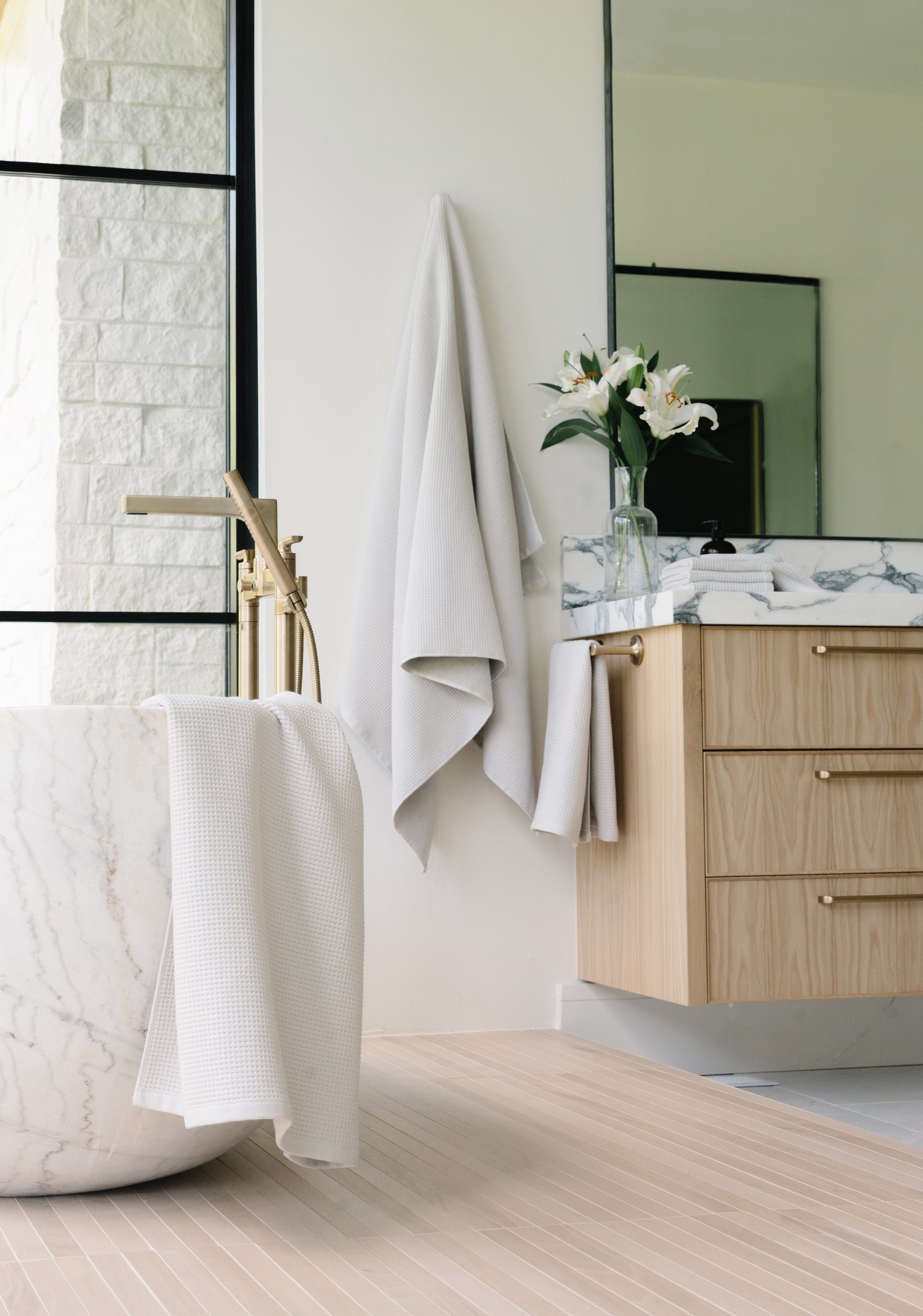 Light Grey Waffle Bath Sheet. The Waffle Bath Sheet in the color Light Grey is photographed in the bathroom. Photo of Light Grey Waffle Bath Towel taken hanging from a towel hook. The towel hook is not seen. A bath tub with another waffle towel draped over the tub is seen in the photo.