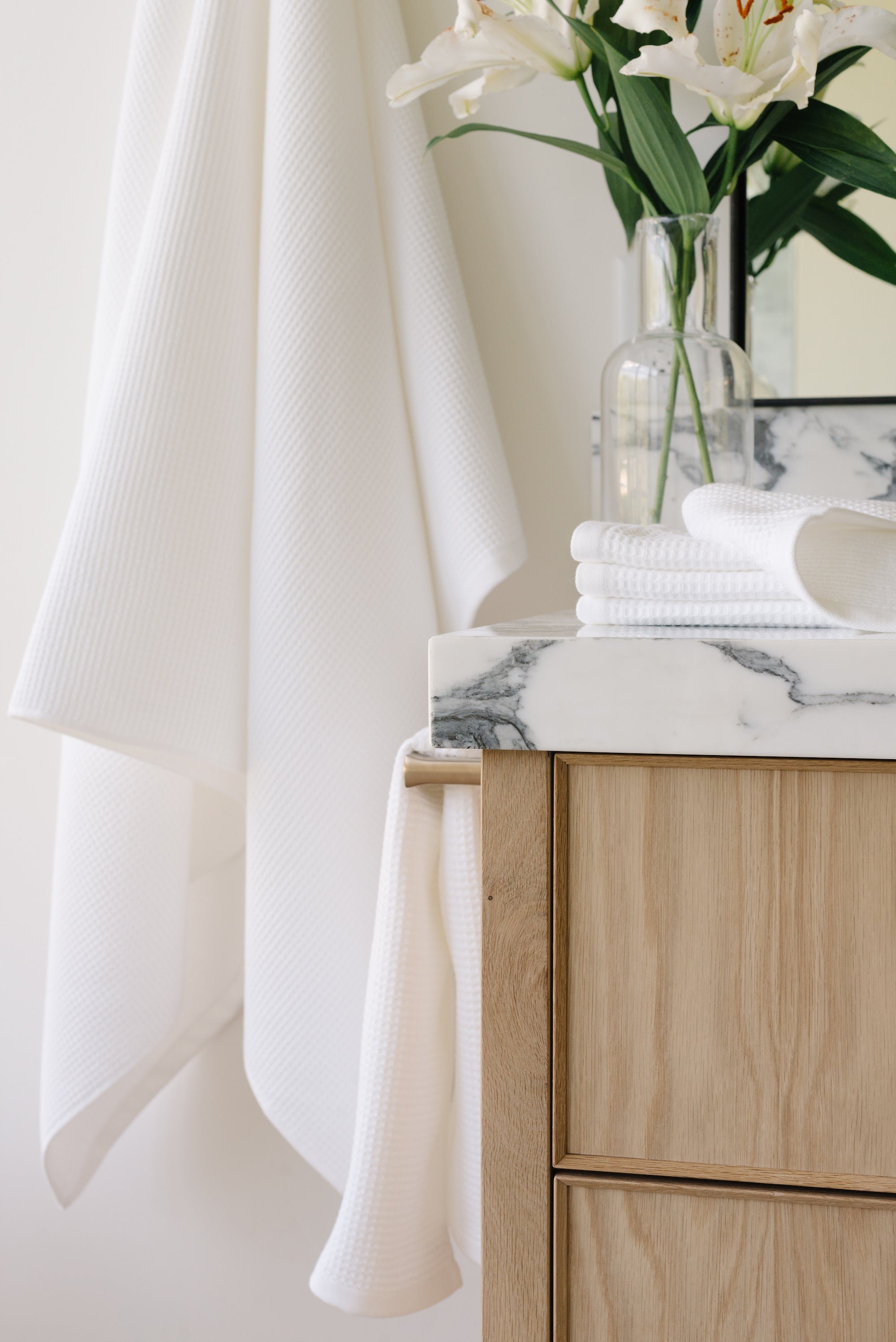 White Waffle Hand Towel hanging from a gold towel bar. The photo was taken at a white marble bathroom sink. A Waffle Bath Towel is seen hanging on the wall in the background. 