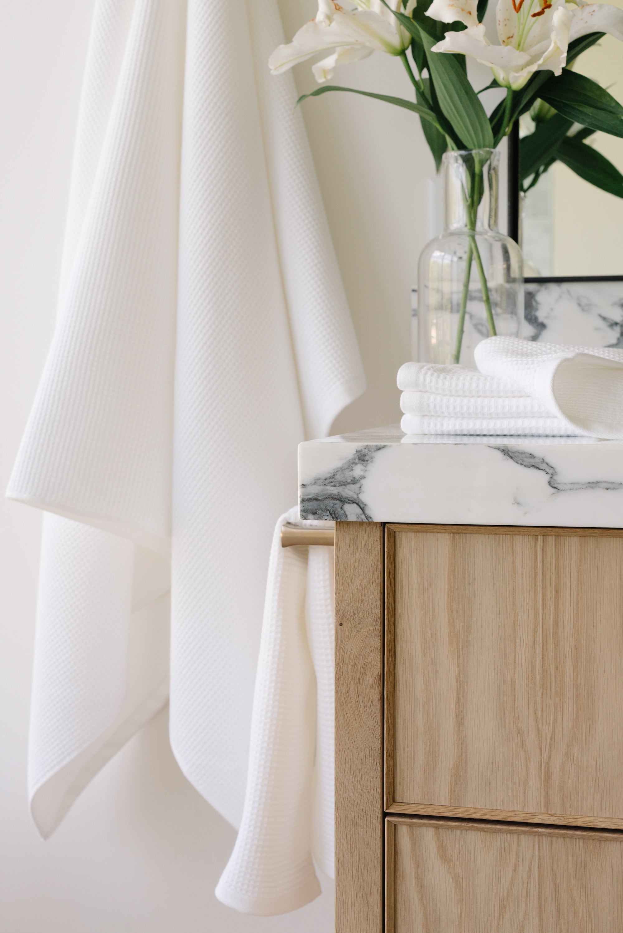 White Waffle Hand Towel hanging from a gold towel bar. The photo was taken at a white marble bathroom sink. A Waffle Bath Towel is seen hanging on the wall in the background. |Color:White