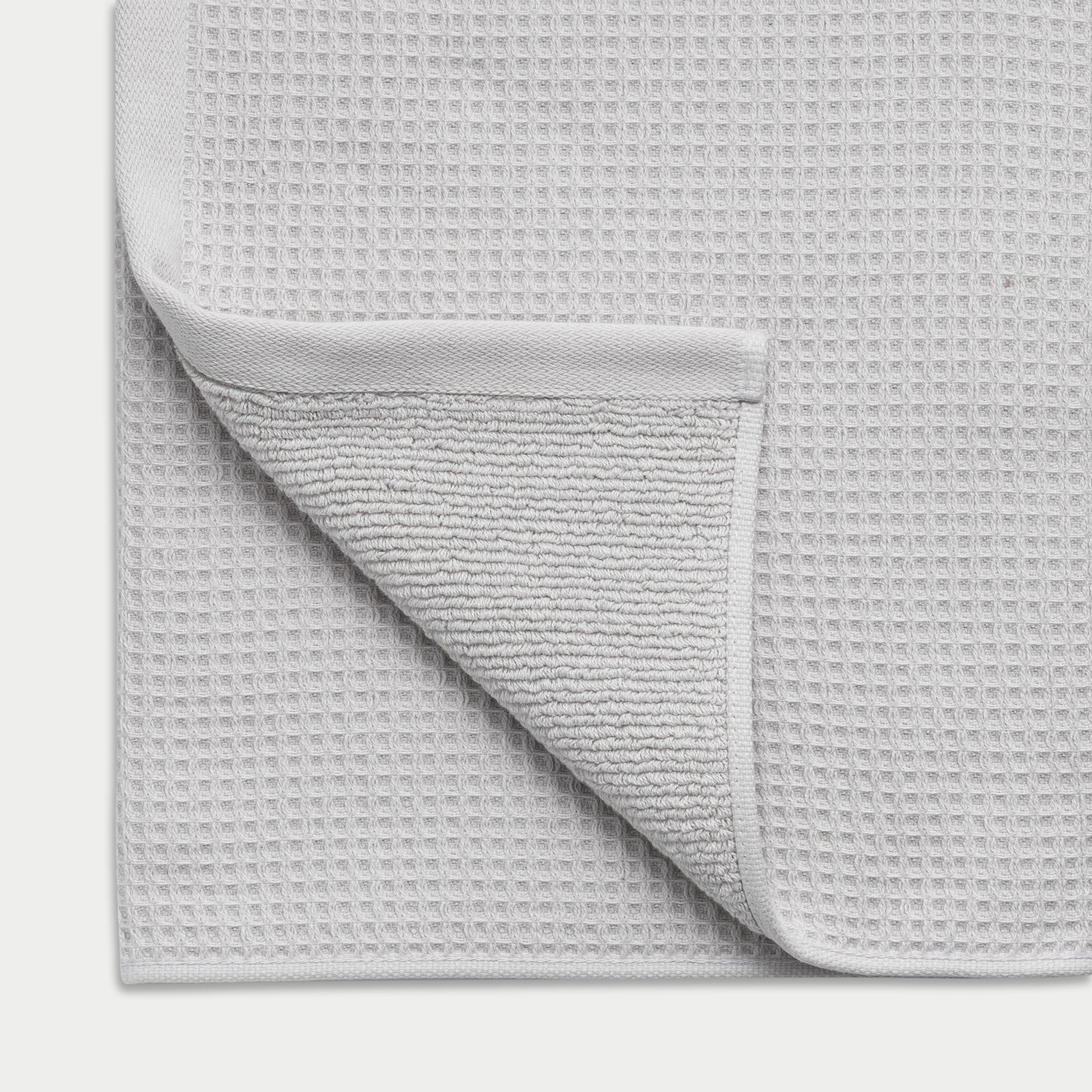 Light Grey Waffle Hand Towel resting flat on a white background. The corner is folded so that both sides of the towel can be seen. 