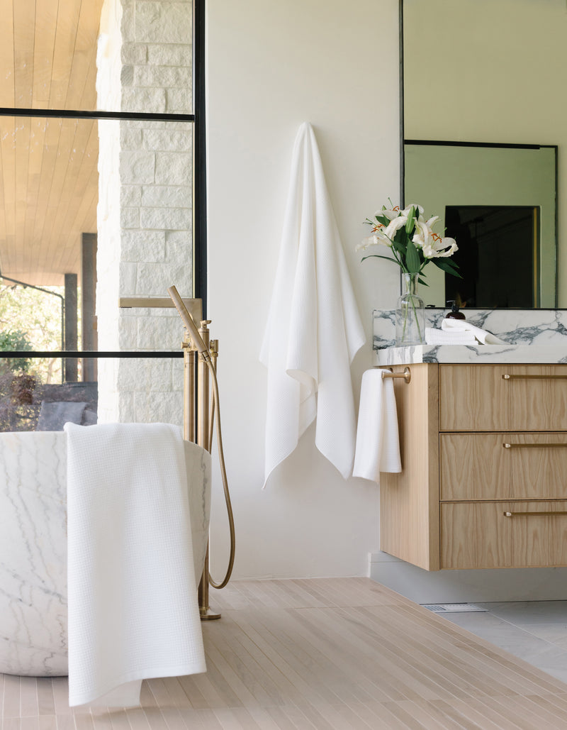 White Waffle Bath Sheet. The Waffle Bath Sheet in the color white is photographed in the bathroom. Photo of Waffle Bath Towel taken hanging from a towel hook. The towel hook is not seen. A bath tub with another waffle towel draped over the tub is seen in the photo.