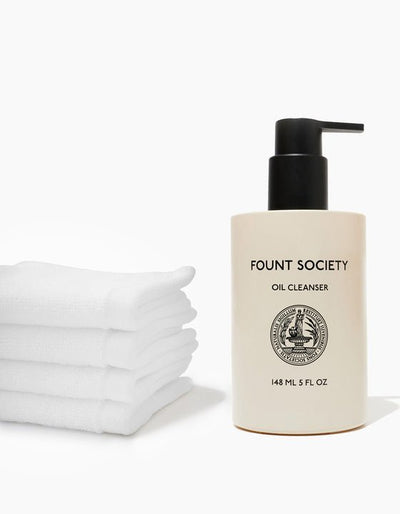 A bottle of Cozy Earth's Revitalizing Pair oil cleanser with a pump stands next to a neatly folded stack of white towels. The bottle is labeled "148 ML 5 FL OZ" and features a minimalistic design with a black cap and text. The background is white.