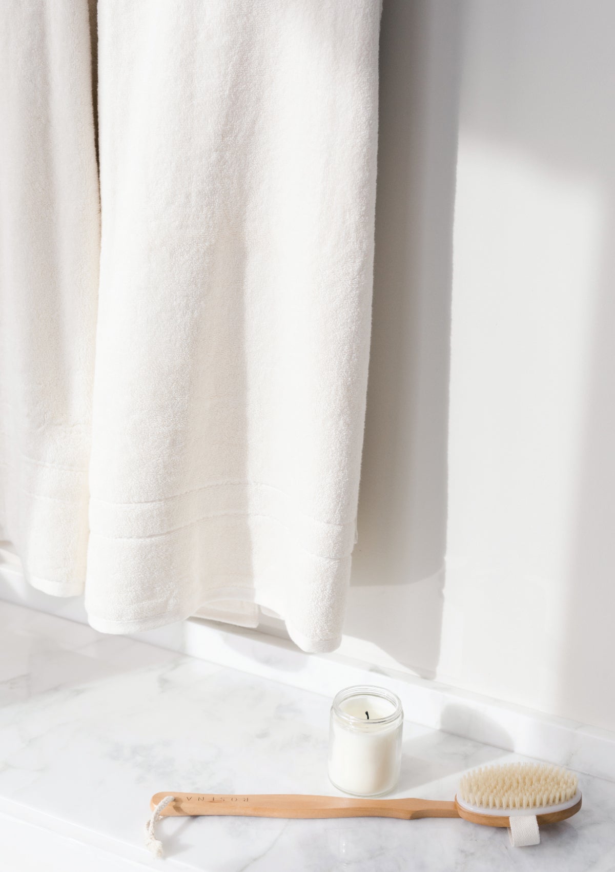 Premium Plush Bath Towels in the color Creme. Photo of Premium Plush Bath Towels taken as a close up of the towel. The photo was taken in a bathroom. There is a scrub brush and a candle on the bathroom floor. 