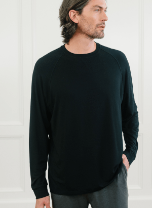 A man with shoulder-length hair is standing and looking to his right. He is wearing a plain black Men's Ultra-Soft Bamboo Pullover Crew from Cozy Earth and dark pants. His right hand is relaxed by his side, and his left hand is in his pocket. The background features a plain white wall with molding.