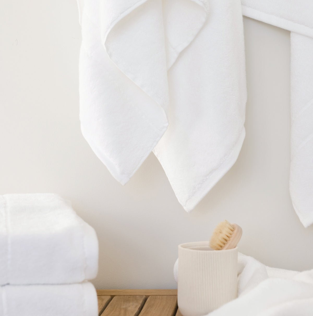 Premium Plush Bath Towels in the color white. Photo of Premium Plush Bath Towels taken in a bathroom showing the towels which are hung from a towel rack. The towels are hung over a wooden shower bench