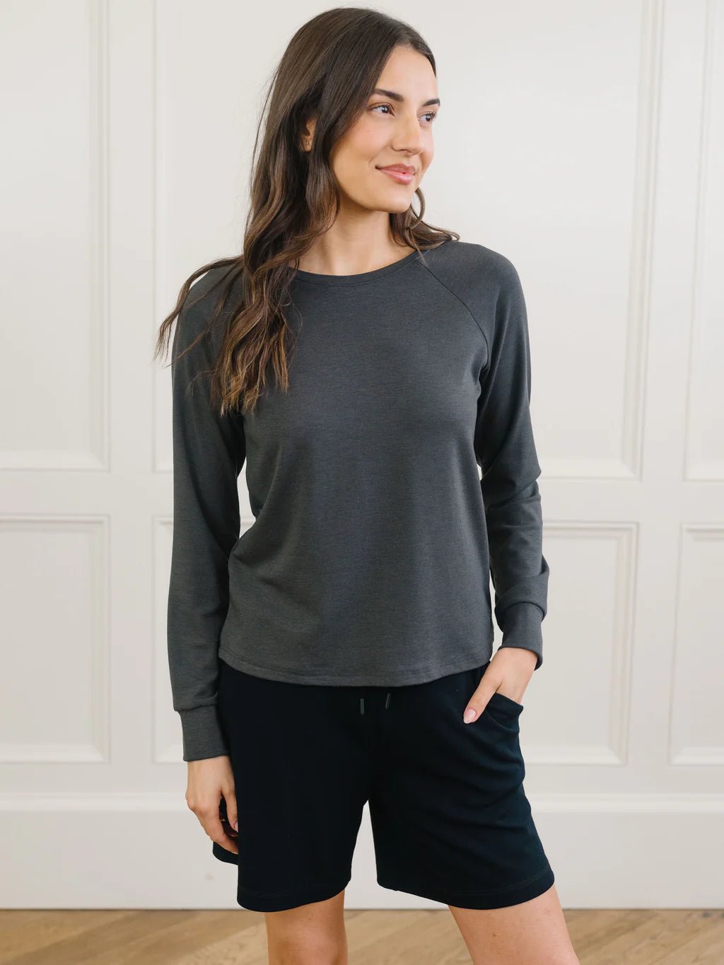 Woman in a grey long-sleeve top and black shorts stands in a room with white paneled walls.
