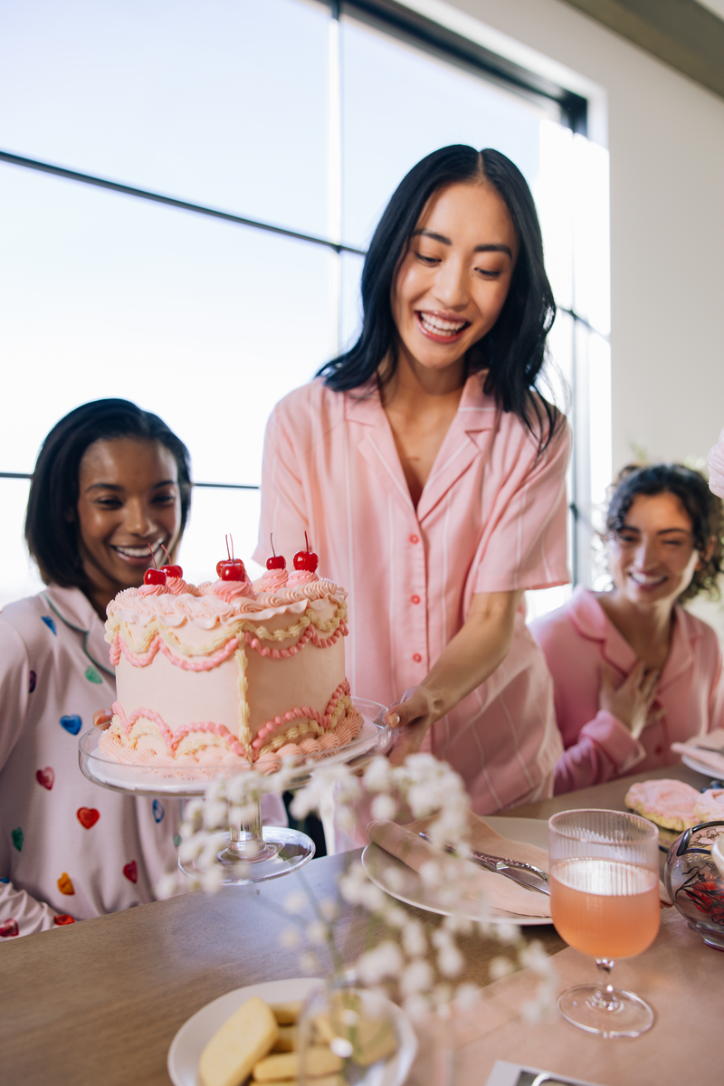 Three people in pajamas celebrating with a decorated pink cake, smiling at a table.