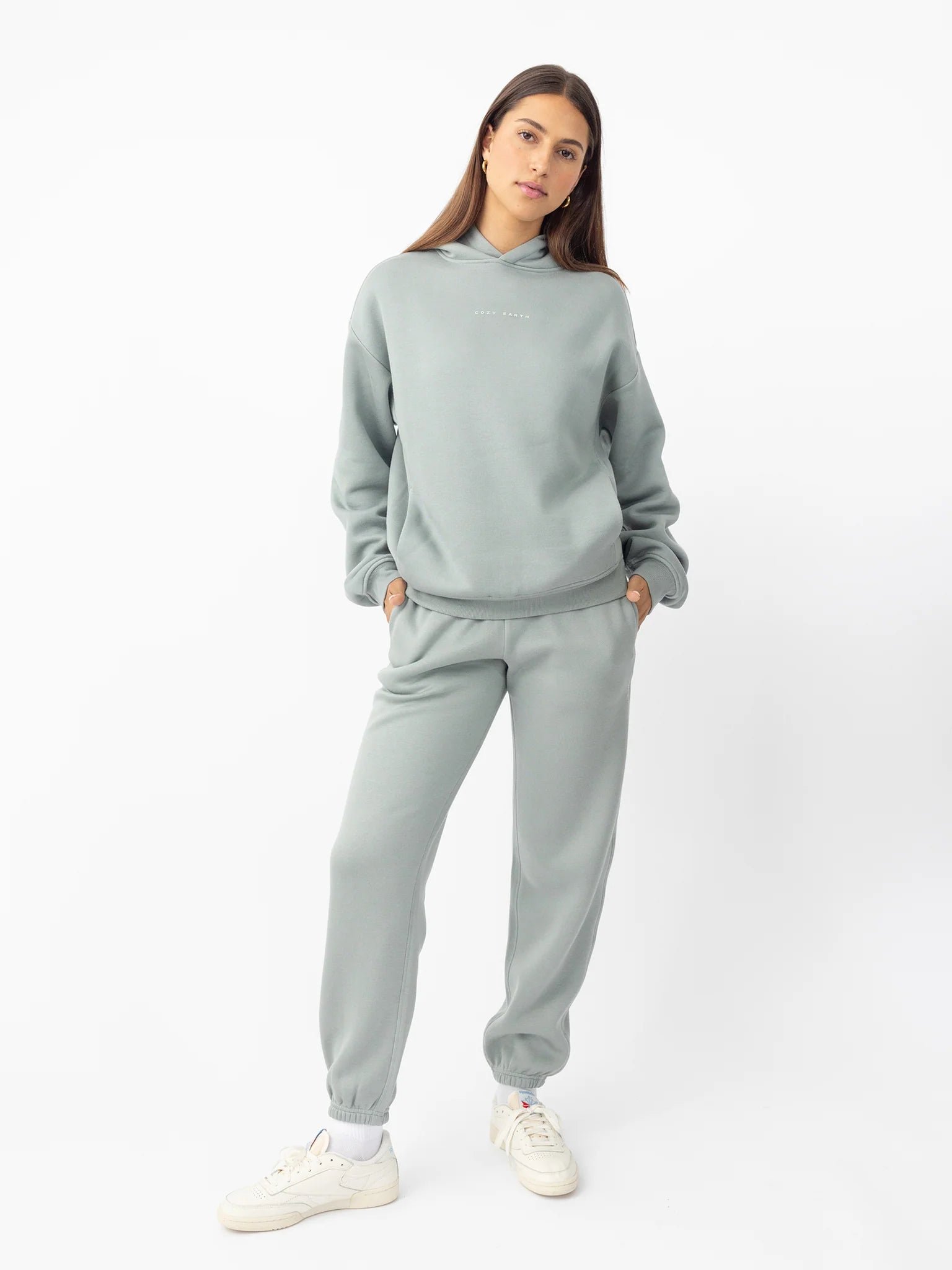 Woman wearing a light green hoodie and sweatpants, posing with hands in pockets.