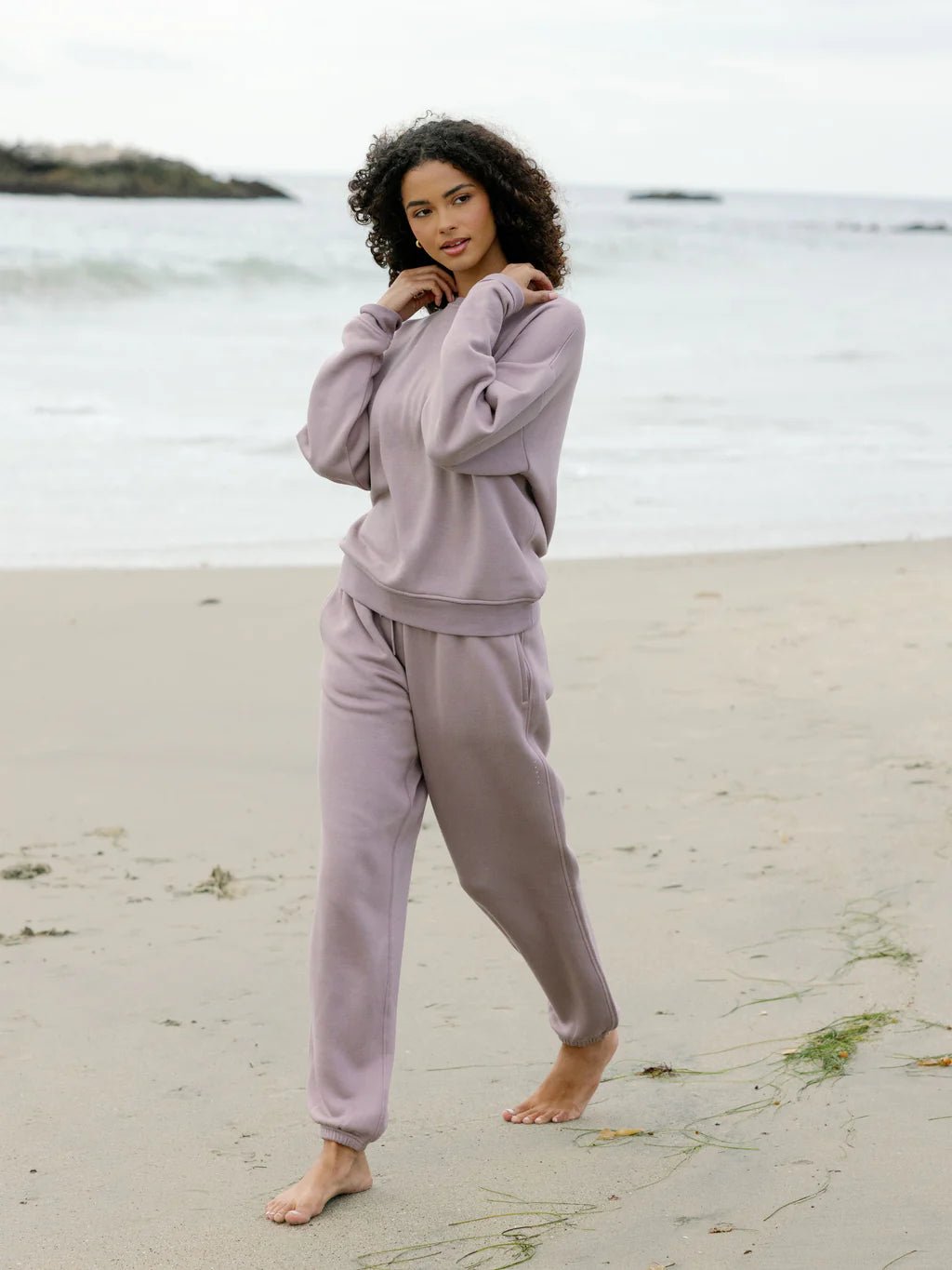 Person in a lavender tracksuit posing barefoot on a beach.