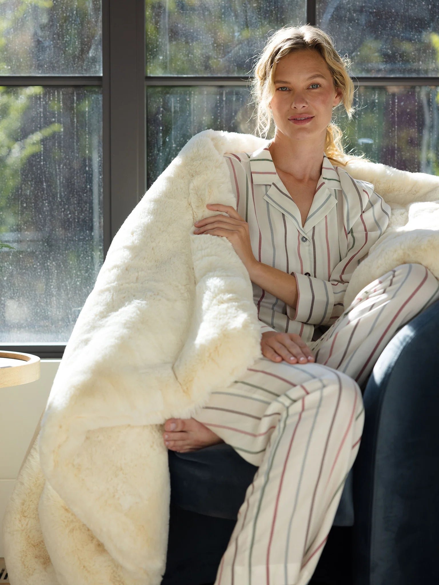 Person in striped pajamas and fluffy blanket sitting by a window.