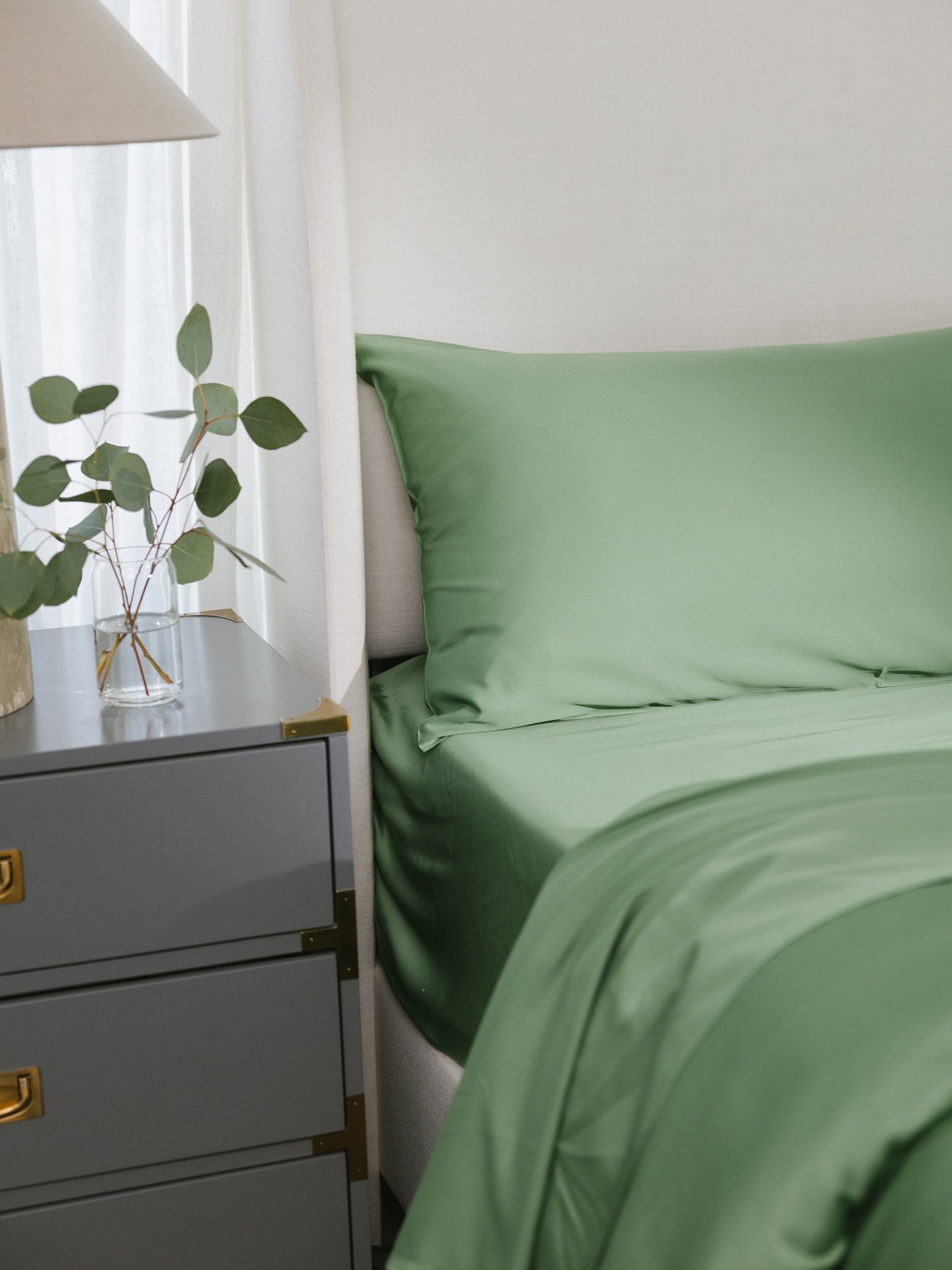 A neatly made bed with green sheets beside a gray nightstand with a plant and lamp.
