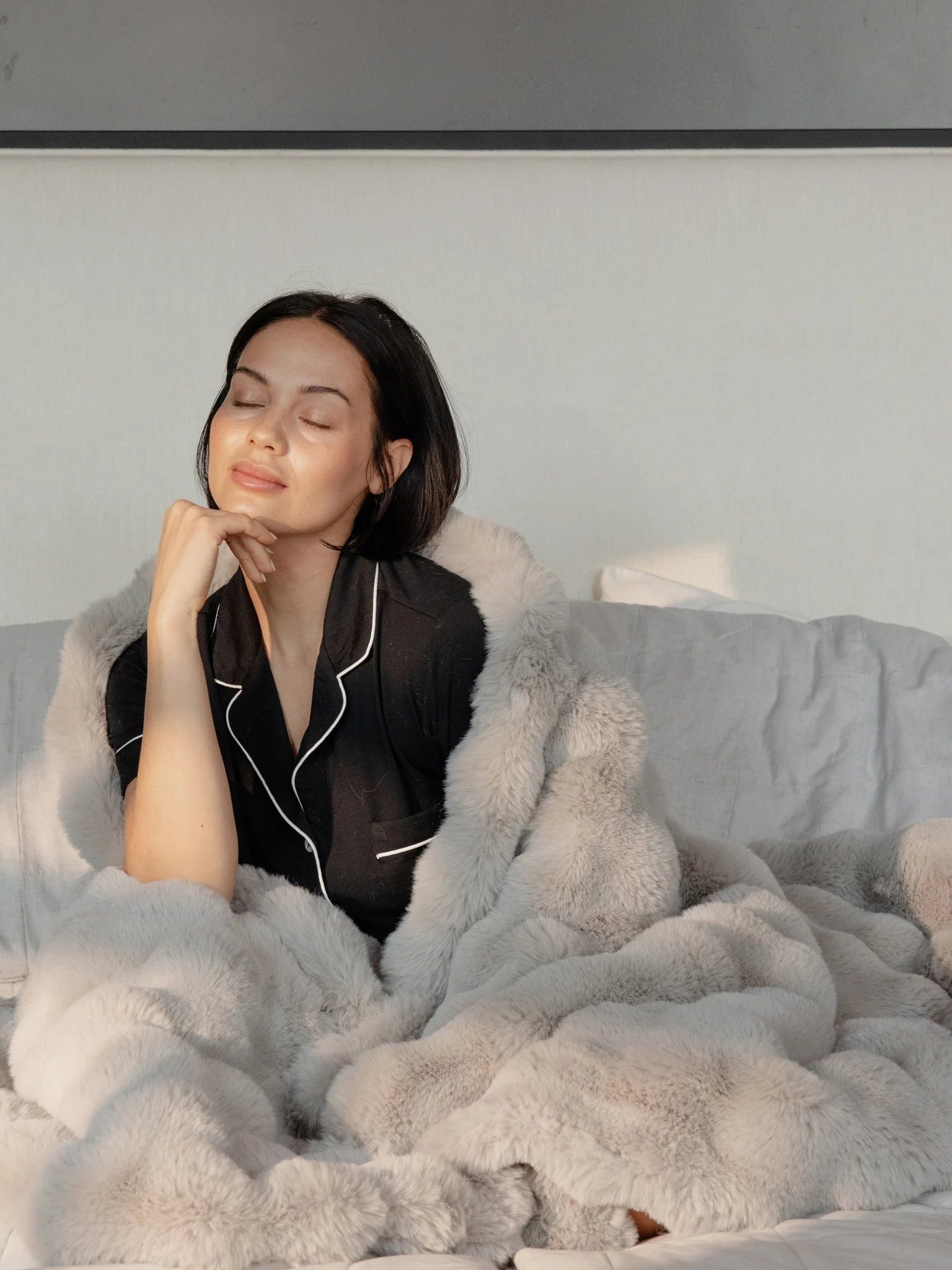Person relaxing with eyes closed, wrapped in a light-colored plush blanket on a bed.