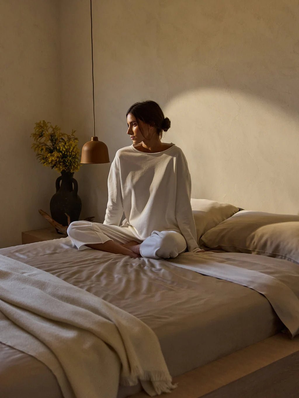 Woman sitting on a bed in a sunlit room, wearing a white outfit.