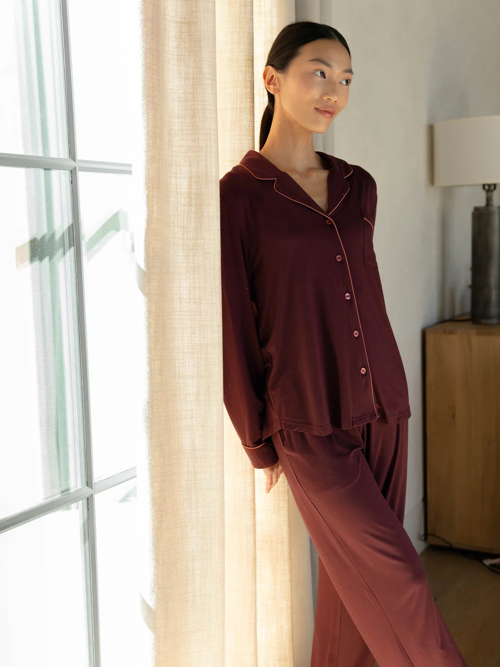 Woman in burgundy pajamas standing by a window with soft lighting.
