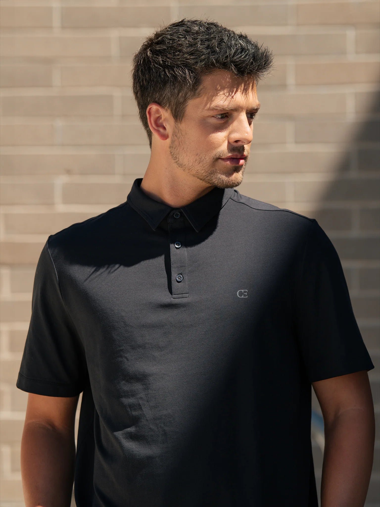 Man wearing a black polo shirt in front of a brick wall.