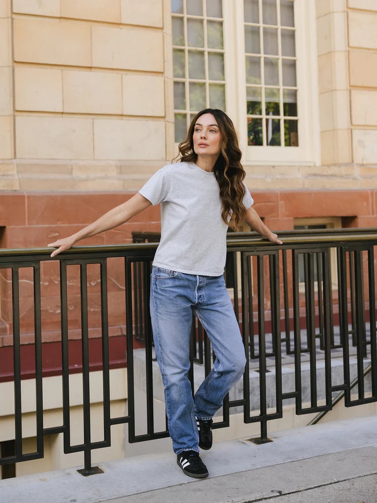 Woman wearing a gray T-shirt and blue jeans, leaning against a black fence outside a building.
