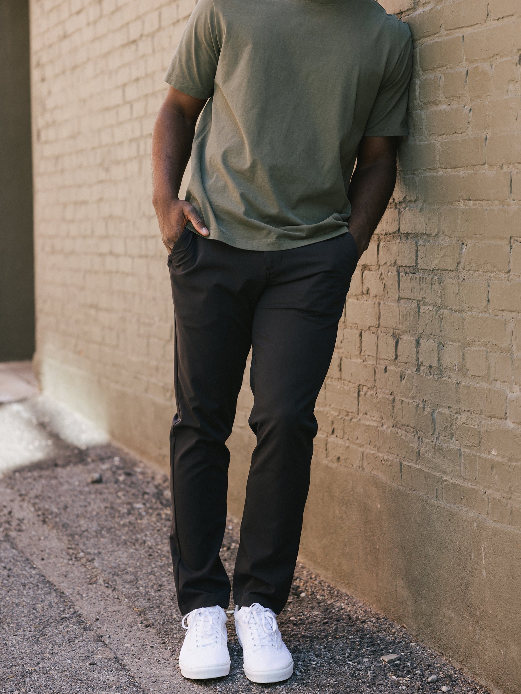 Person in a green t-shirt and black pants leaning against a brick wall.