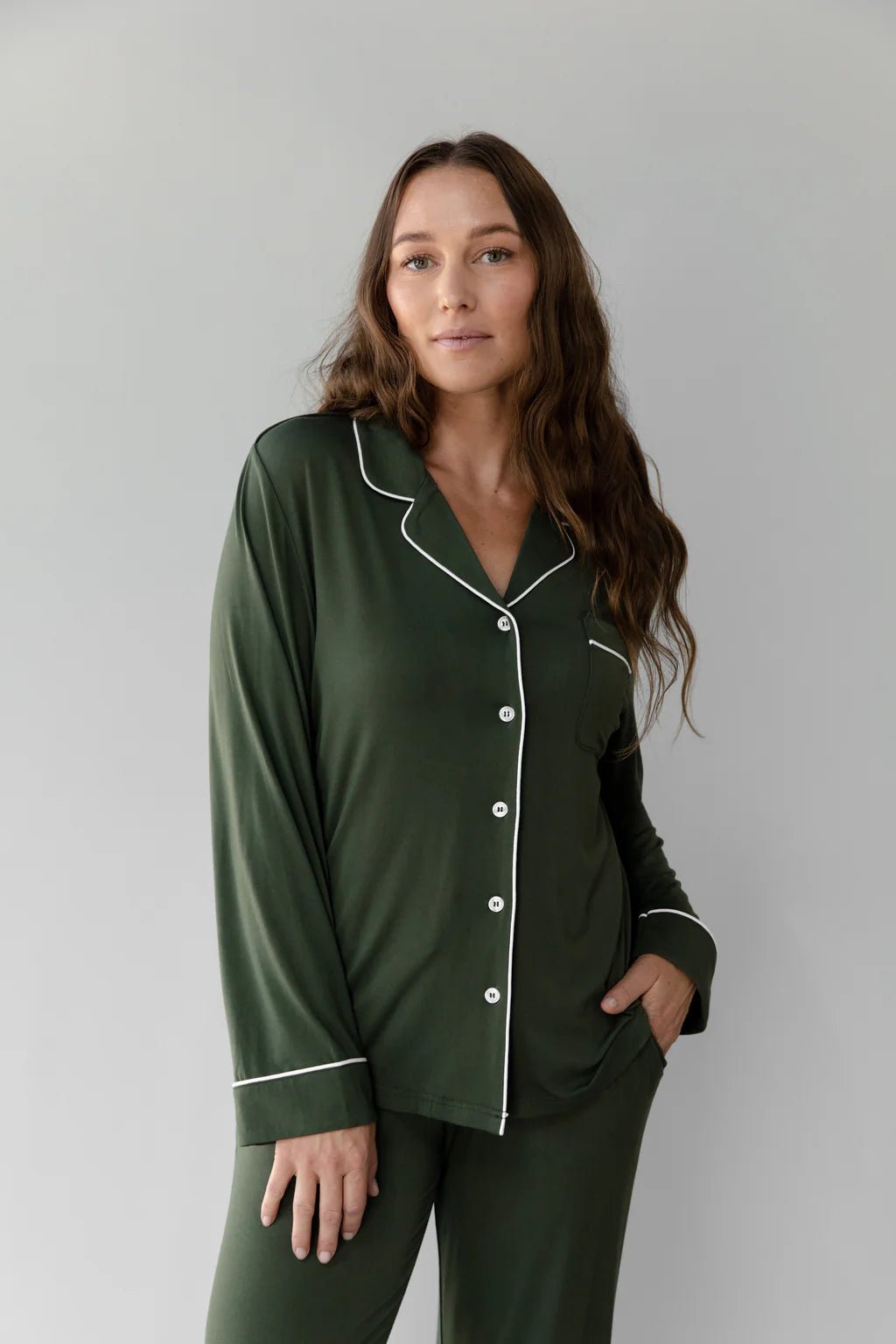 Woman wearing dark green pajamas with white piping, standing against a neutral background.