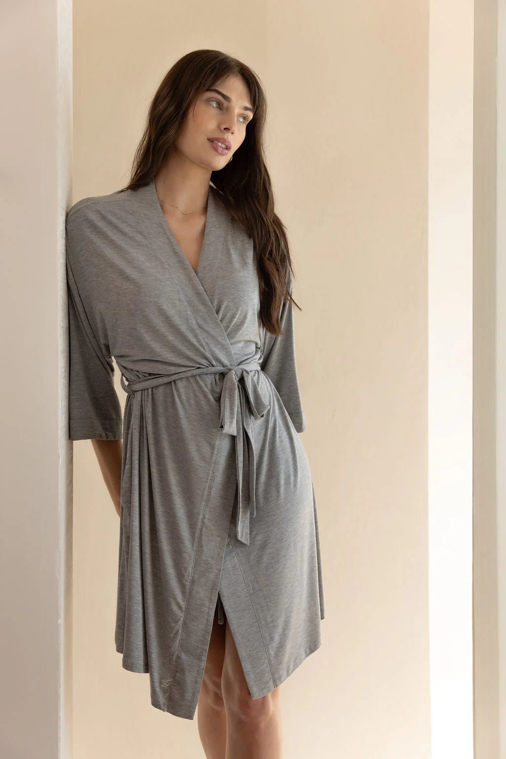 A person wearing a light grey wrap-around dress standing against a wall.