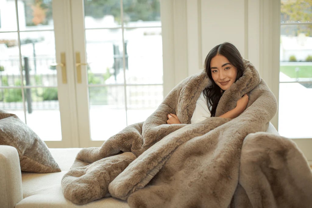 Person wrapped in a fluffy, brown blanket on a cozy couch by a window.