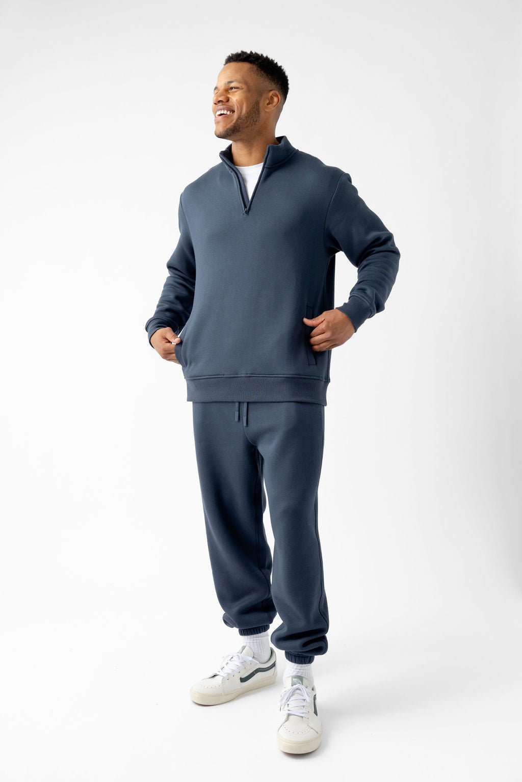 Man wearing a blue tracksuit and white sneakers, smiling and standing against a plain background.