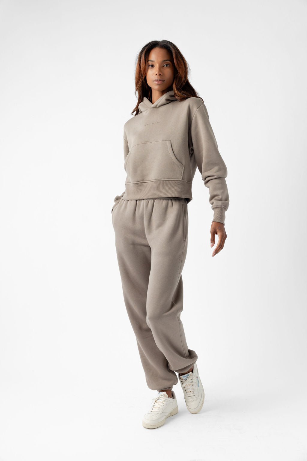 Person wearing a beige hoodie and matching sweatpants with white sneakers against a plain background.