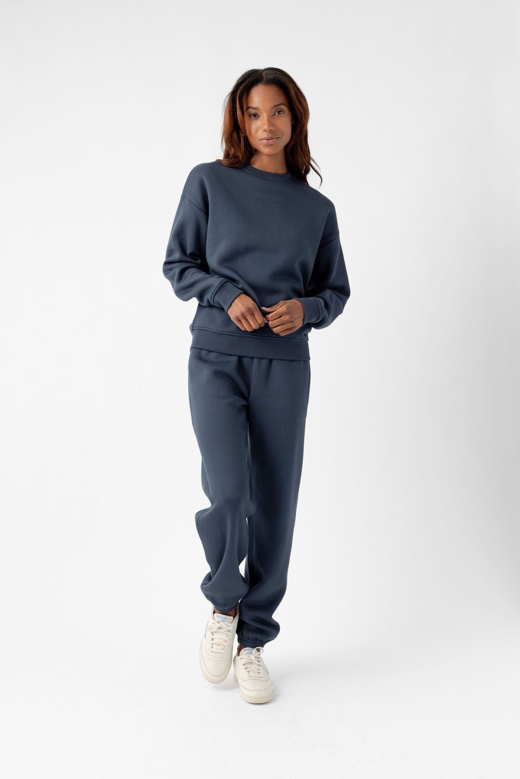 Person standing and wearing a navy sweatsuit and white sneakers against a plain background.