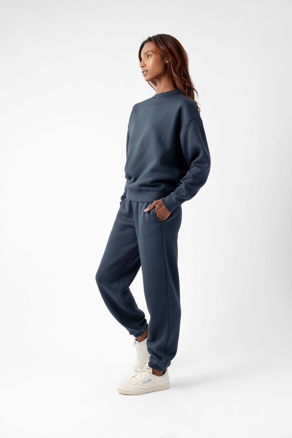 Person wearing a dark colored sweatshirt, matching sweatpants, and white sneakers, standing against a white background.