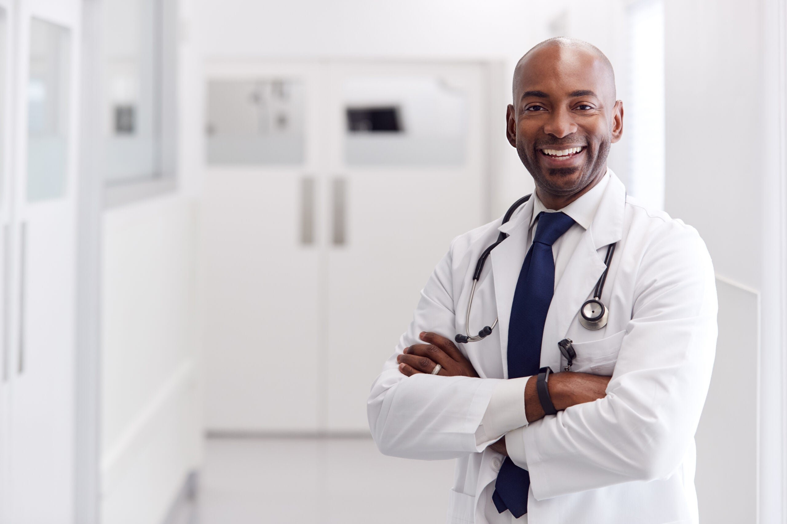 Smiling doctor in a white coat with a stethoscope, standing with arms crossed in a hospital corridor.
