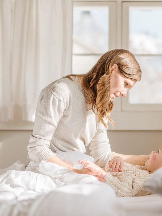 Woman with beige top playing with child on a bed.